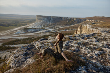 Woman sitting on cliff overlooking valley with mountains in background on adventure trip to explore...