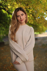 A beautiful fashionable young blonde girl stands in an autumn park on leaves in a gray dress and sneakers