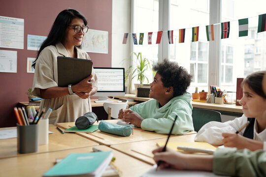 Young smiling teacher with tablet looking at clever schoolboy and answering his question at lesson of English grammar