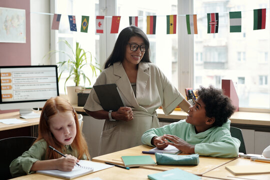 Adorable schoolboy sitting by desk and talking to young smiling female teacher of English grammar in front of schoolgirl