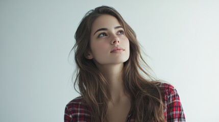 Fototapeta premium A young woman with long brown hair, wearing a red and black plaid shirt, gazes thoughtfully upward, lost in contemplation. Her expression is serene and introspective.
