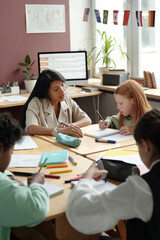 Cute diligent schoolgirl bending over desk and writing down grammar rules in copybook while young teacher checking her notes