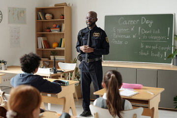 Confident African American policeman in uniform standing in front of class of youthful kids and talking about his job and career