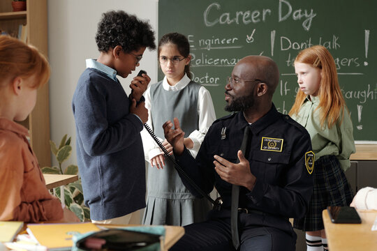 Cute African American schoolboy speaking in walkie-talkie at lesson while standing in front of policeman giving instructions - Powered by Adobe