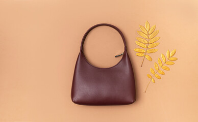 Women's leather elegant fashion baguette bag in burgundy color with golden autumn leaves on brown background. Top view with copy space. Autumn female fashion accessory