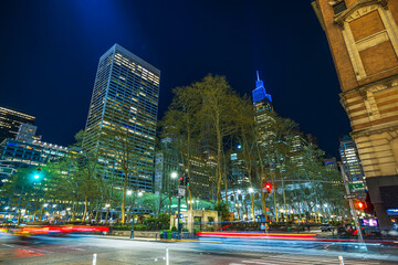 Night view of Bryant Park in Manhattan with city lights, tall buildings, and light trails from passing car headlights.