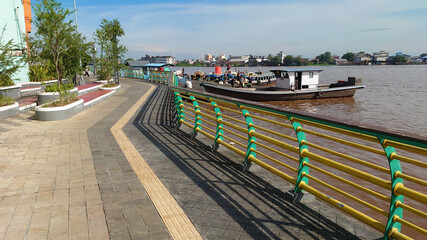 A peaceful riverside walkway features vibrant railings, lush greenery, and moored boats, under a clear blue sky