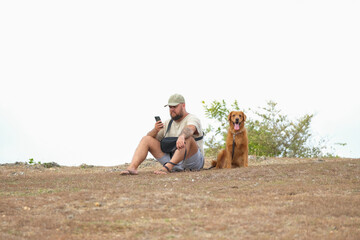 A young man in a cap sits on the grass in a park and looks at his phone. A golden retriever sits next to him, sticking out its tongue and looking into the distance. Walking with a dog in summer.