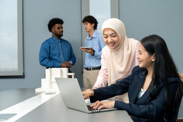 Two professionals working together on a laptop, exchanging ideas, while other team members discuss in the background of a modern office.