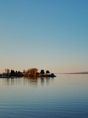 Naklejka premium Tranquil view of the Ottawa River at sunset on a crisp autumn day, with golden light illuminating the trees on a small riverside island, surrounded by calm, reflective water under a clear sky.