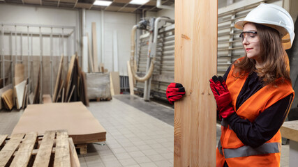 Woman employee furniture workshop. Girl carpenter holds board in hands. Woman worker at manufacturing plant. Girl engaged in woodworking. Carpenter in orange vest and helmet. Furniture business owner