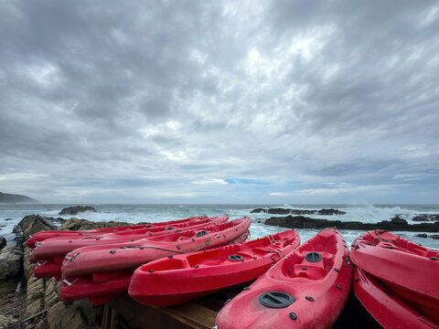 Red kayaks stacked on the coast. Ocean waves crashing against the rocks, cloudy sky. Tsitsikamma national park, South Africa