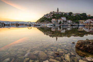 Harbor and beach with an old village on a hill. Boats are reflected in the water, the sun rises on the horizon of the Adriatic. Beautiful landscape shot from Vrbnik, Krk, Croatia