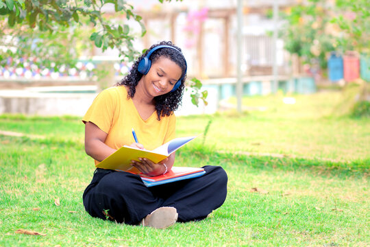 Estudante brasileira negra, sentada na grama de um pra&ccedil;a para estudar. Ela est&aacute; sorrindo enquanto escreve no seu caderno. Estudante se preparando pro vestibular, prova ou enem.  