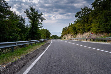 Road landscape forest, mountains and sea on the horizon. Empty long mountain road to the horizon on a sunny summer day with dramatic cloudy sky. Highway turn panoramic view.