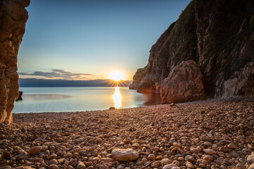 Hidden cove and beach with an old hilltop village. The sunrise is reflected in the water. A beautiful landscape shot framed by cliffs and the Adriatic sea from Vrbnik, Krk, Croatia