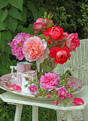 Red and pink roses still life in pretty antique porcelain jugs and vases a plate on a wicker  table, outdoors, in a garden