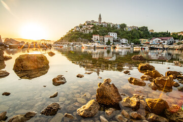 Harbor and beach with an old village on a hill. Boats are reflected in the water, the sun rises on the horizon of the Adriatic. Beautiful landscape shot from Vrbnik, Krk, Croatia