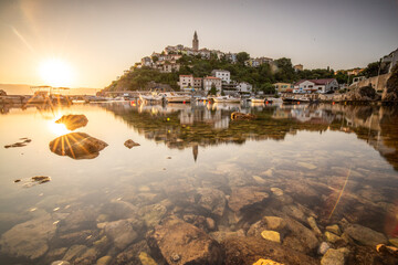 Harbor and beach with an old village on a hill. Boats are reflected in the water, the sun rises on...