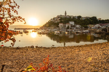 Harbor and beach with an old village on a hill. Boats are reflected in the water, the sun rises on the horizon of the Adriatic. Beautiful landscape shot from Vrbnik, Krk, Croatia