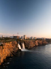 Tranquil coastal landscape at sunrise, featuring a cascading waterfall flowing gently into the calm sea. Warm golden hues of the rising sun reflect on the water
