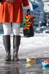 Closeup of woman in a orange coat and rubber boots walking down the street with a bouquet of tulips in spring city