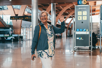 Smiling senior woman travels in airport area with backpack waving hand walking towards board gate....