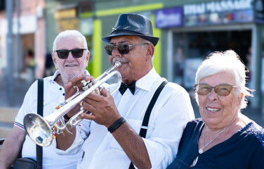 Portrait of jazz artist in open air concert playing among passers by. Old man in black hat and sunglasses playing trumpet in street. © luciano
