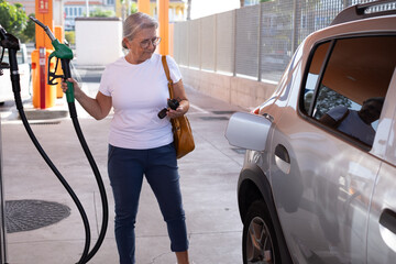 Mature modern woman fills up her car at self-service fuel pump in European gas station - inflation,...