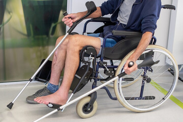 Obraz premium middle-aged man sits in a wheelchair with his bare leg, which has been removed from a plaster cast, waiting for an X-ray outside a doctor's office in a clinic, holding crutches in his hands,