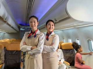 Two flight attendants or air hostess smiling confidently in beige uniform standing with arms crossed in airplane cabin. Passengers child sitting by window with mother. Airline transportation and touri