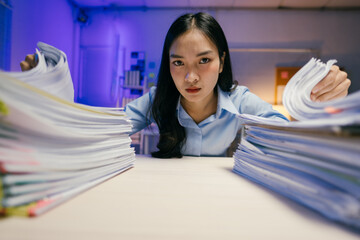Young asian businesswoman looking stressed while working overtime at night in office with huge piles of paperwork on her desk, struggling with burnout and workload