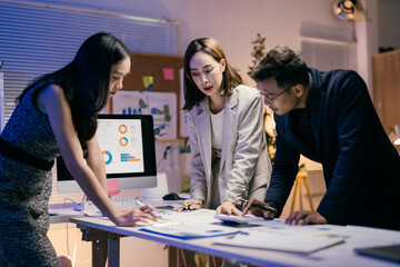Obraz premium Three asian businesspeople working together late at night in office meeting room analyzing financial charts displayed on a desktop computer and printed on paper documents