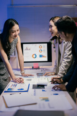 Three asian businesspeople are examining financial reports and discussing data displayed on a computer screen, working late in a modern office