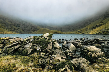 A body of water nestled between a foggy mountain range. The water is calm and reflects the cloudy sky. Rocks and grass are scattered along the shore.