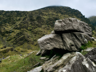 A striking granite boulder rests firmly on a verdant hillside surrounded by rugged terrain. The scene is set against a backdrop of cloudy skies, highlighting the natural beauty.