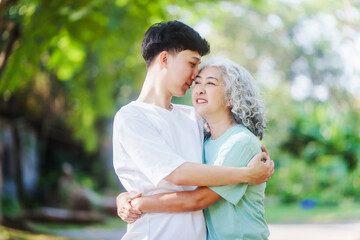 Fototapeta premium Young son and his mature mom asian people share loving hug in beautiful park, surrounded by nature. Their happiness radiates as they bond outdoors, celebrating family love and togetherness.