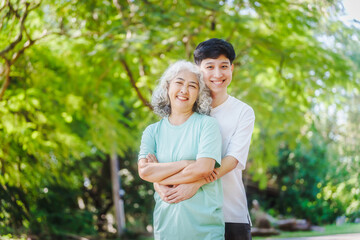 Fototapeta premium Young son and his mature mom asian people share loving hug in beautiful park, surrounded by nature. Their happiness radiates as they bond outdoors, celebrating family love and togetherness.