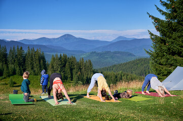 Group of people doing yoga pose outdoor in camping in the mountains. Adults and children on yoga mats, each doing yoga pose under clear blue sky in morning.