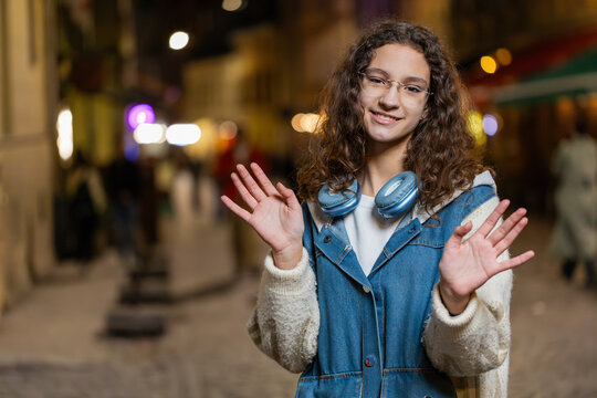 Hello. Teenage girl in casual clothes smiling friendly at camera, waving hands gesturing hi, greeting or goodbye, welcoming with hospitable expression. Happy girl on urban city street at night outside
