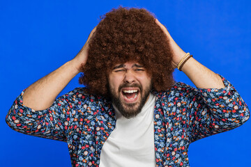 Irritated frustrated man with Afro hairstyle wig screams from stress tension problems feels horror hopelessness fear surprise shock expresses rage nervous, quarrel. Young guy on blue wall background