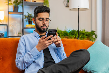 Happy young man in casual clothes thinking and text messaging on smartphone. Indian guy sitting on...