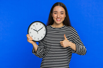 It is your time, hurry up. Caucasian woman showing time on wall office clock, ok, thumb up, approve, pointing finger at camera, advertisement, deadline. Young girl isolated on studio blue background