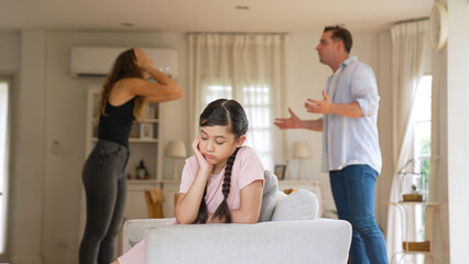 Annoyed and unhappy young girl sitting on sofa trapped in middle of tension by her parent argument...