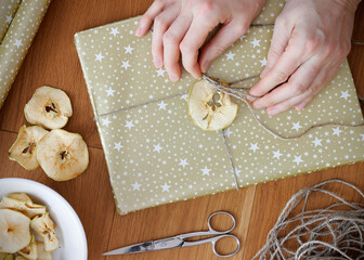 Female hands wrapping Christmas present. Christmas gift wrapped in gold paper and decorated with dried apple, dried apples in a bowl, ribbon and scissors on wooden floor. Christmas flat lay. © KatuSka