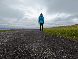 Rear view of a single female standing on a winding gravel road. She is dressed casually and takes in the beautiful views of nature. Grassy field surrounds the road. Nólsoy, Faroe Islands.