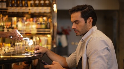 Smart man drinking cocktail in Old fashioned waiting friends or couple while chatting on smartphone to wait at counter bar with bartender preparing drinks at nightclub Friday meeting night. Vinosity.