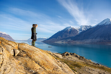 Man photographs the fjords and mountains of Greenland, with schooner anchored in the deep blue waters. Forsbladfjorden, Northeast Greenland National Park.