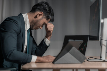 Stressed businessman watches stock market fall on laptop with falling stock graph. A businessman in a suit sits at a table with his hands covering his face. Stock market crash on computer screen