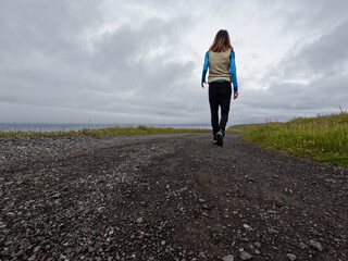 Rear view of a single female walking on a gravel road overlooking the sea. She is dressed casually and takes in the beautiful views of nature. Nólsoy, Faroe Islands.